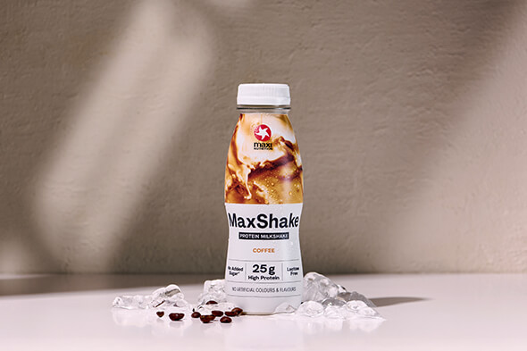 MaxiNutrition MaxShake Coffee vor einer braunen Wand mit Eiswürfeln und Kaffeebohnen neben der MaxShake Flasche.