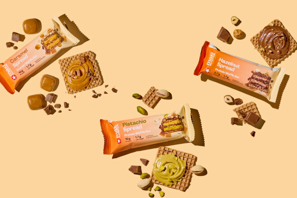 maxinutrition protein waffle bar pistachio spread, caramel spread und hazelnut spread auf beigem hintergrund mit zutaten der einzelnen riegel