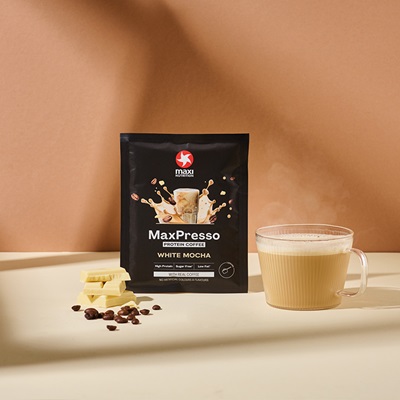 maxinutrition_MaxPresso_white-mocha_Sachet_stehend_vor_brauner_wand_neben_Kaffeebohnen_mit_warmem_shake_in_einer_tasse